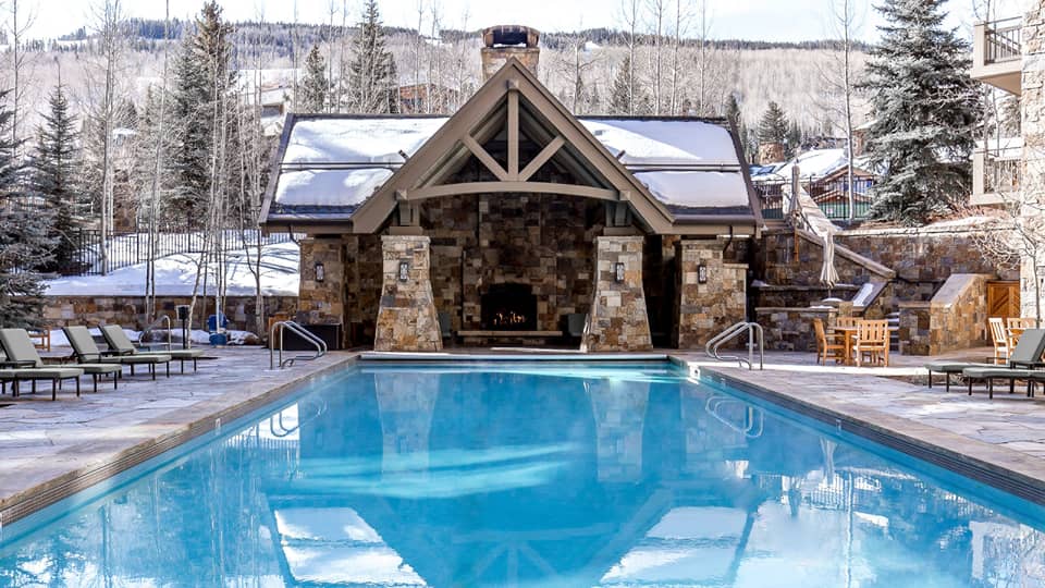Outdoor pool with lounge chairs, a cabin with a fireplace and pine trees in the background.