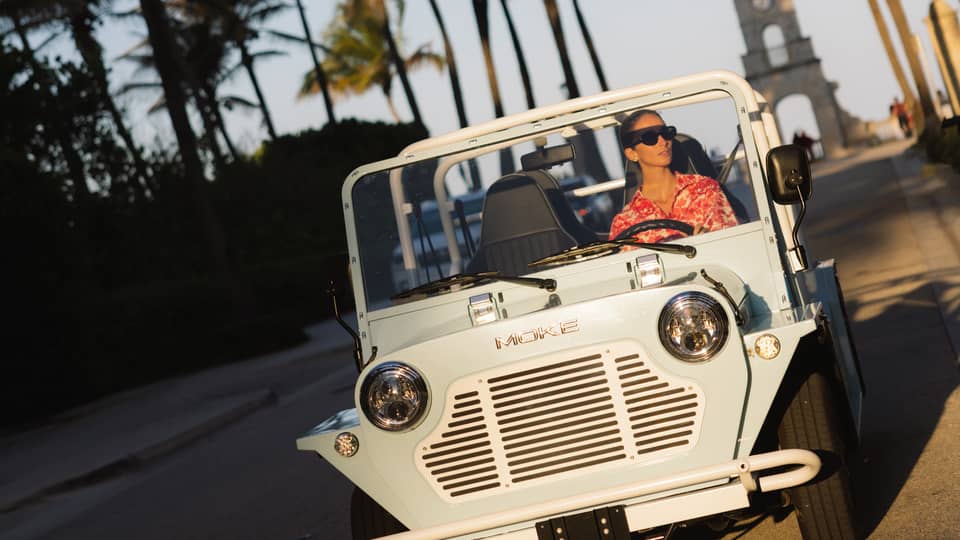 Guest wearing a red-and-white tropical shirt rides in a white MOKE along a palm tree?lined street at dusk