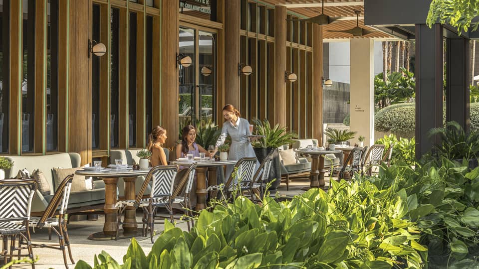Two dining patrons are served by a waiter on the shaded patio of a restaurant