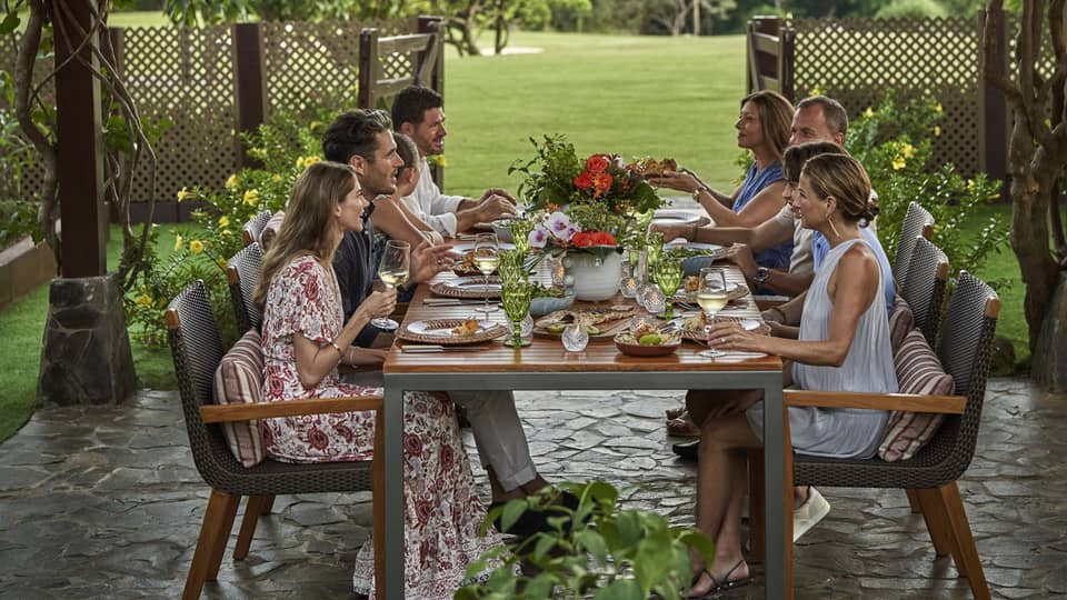 Eight people sit around a long dining table set up outside beneath a canopy of greenery and string lights