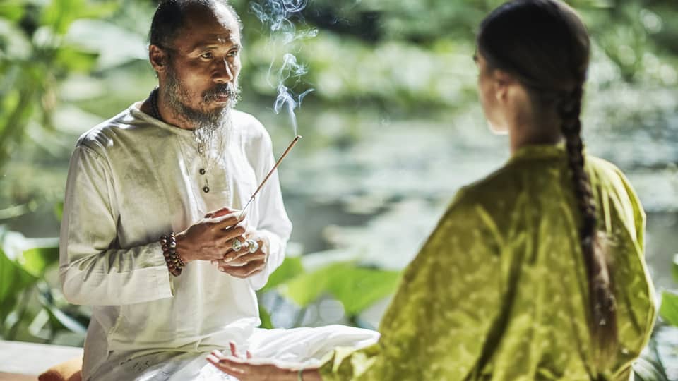 Set against a backdrop of greenery, Djik Dewa holds a stick of burning incense before a meditative cross-legged guest. 