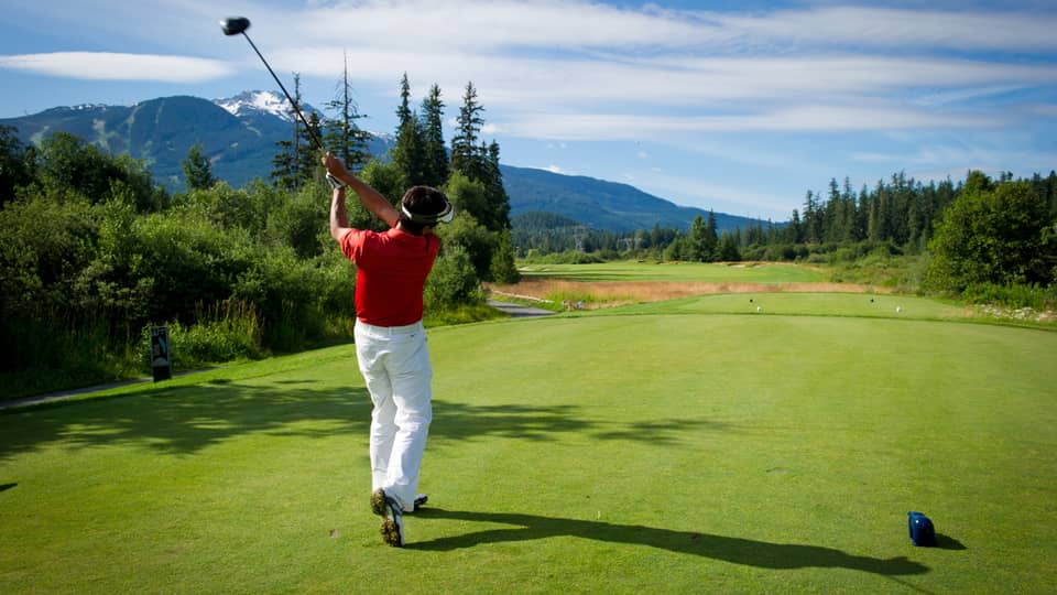 Back view of man swinging golf club on green, mountains in distance 