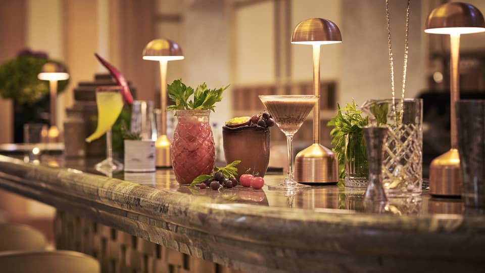 Cocktails in various glassware lined up on bar under copper desk lights