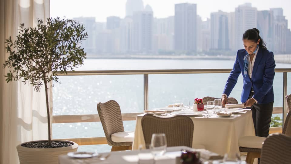 A four seasons staff setting an indoor  Cafe Milano table overlooking the water 