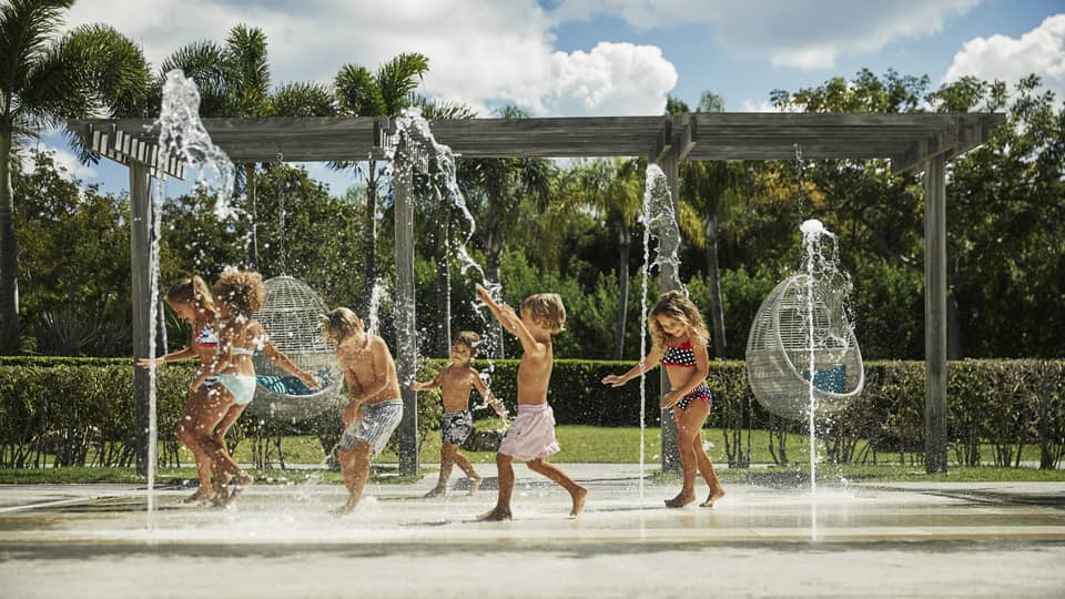 Kids in swimsuits laugh and run under outdoor splash pad, fountain