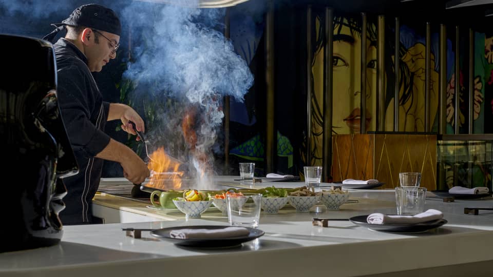 Chef in black coat cooks dish over open flame in show kitchen