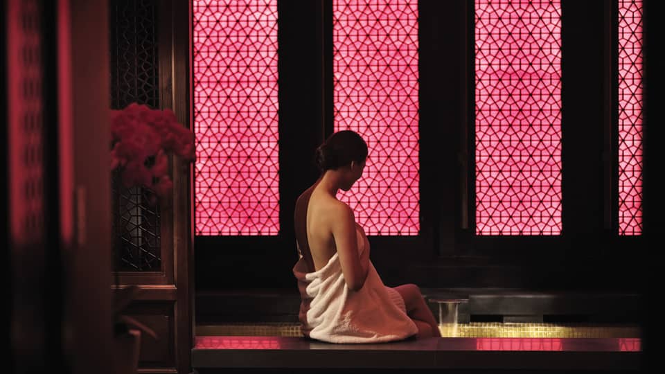 Woman wearing white towel sits on edge of dark spa tub by wall with pink lights
