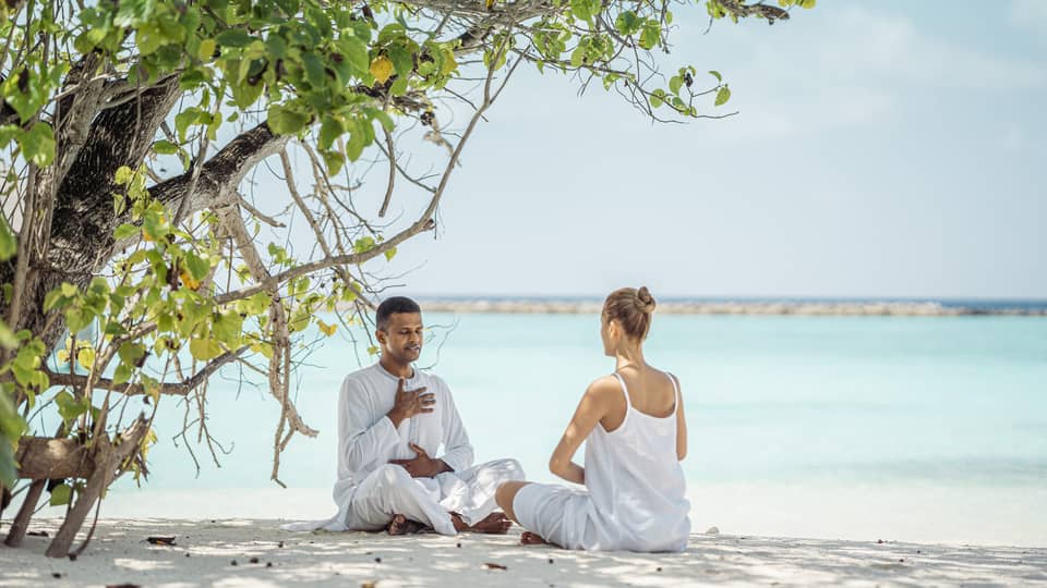 Dr. Arun K. Tomson practises breathing exercises with female guest on the beach under the shade of a tree