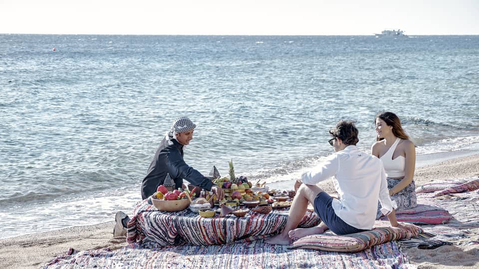 Three people sit on a beach around a low table set with a luxurious array of fruit, bread, and clay dishes full of food.