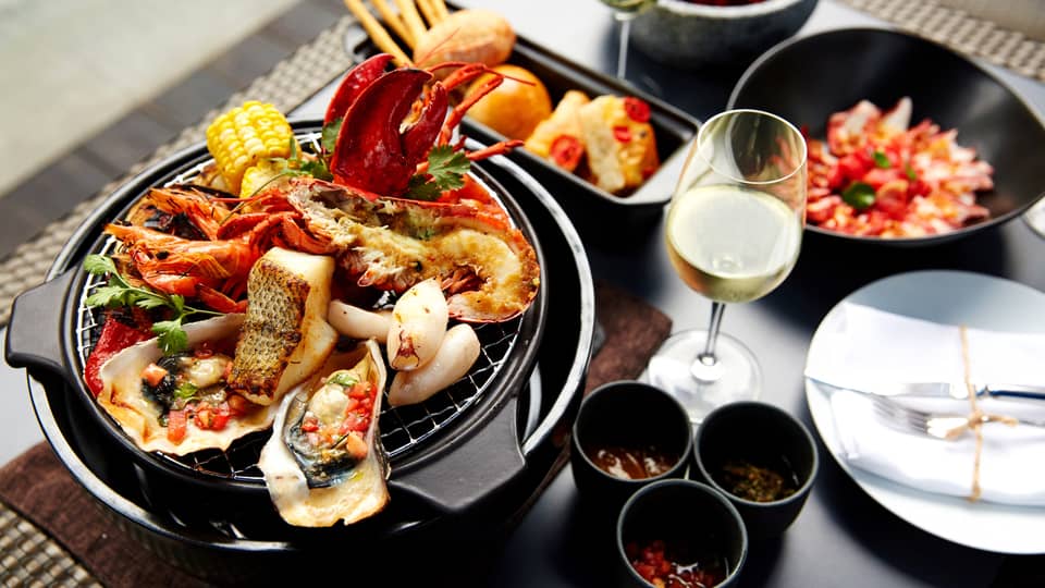 Grilled fish, oysters, lobster, crab on grill plate next to white wine, garnishes