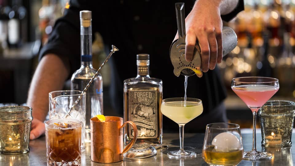 Various cocktail glasses and liquor bottles on bar as mixologist pours martini out of shaker