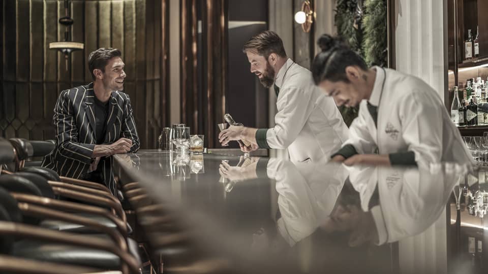 Bartender preparing a drink while a customer sits at the bar, both smiling, in a modern, upscale bar with a reflective countertop and warm lighting.