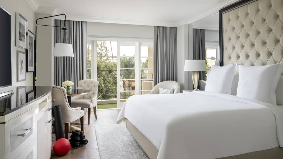 A hotel room with large open window, bed with white sheets, dresser, two chairs, an exercise ball and weights.