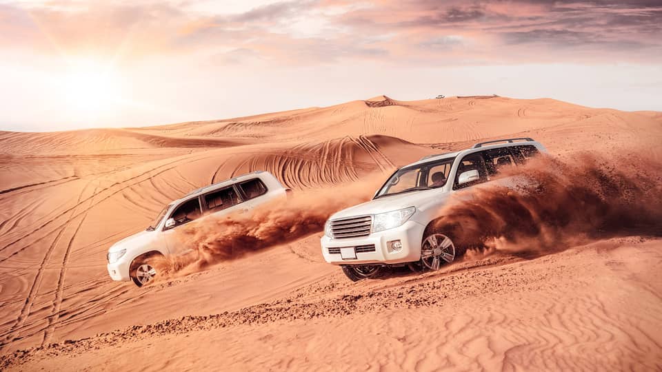 Sand flies from tires of two white SUV's driving across dunes