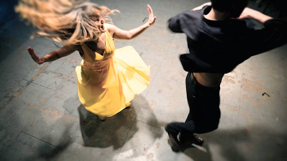 Woman in a yellow dress is spinning on the dance floor as a man wearing all black dances next to her