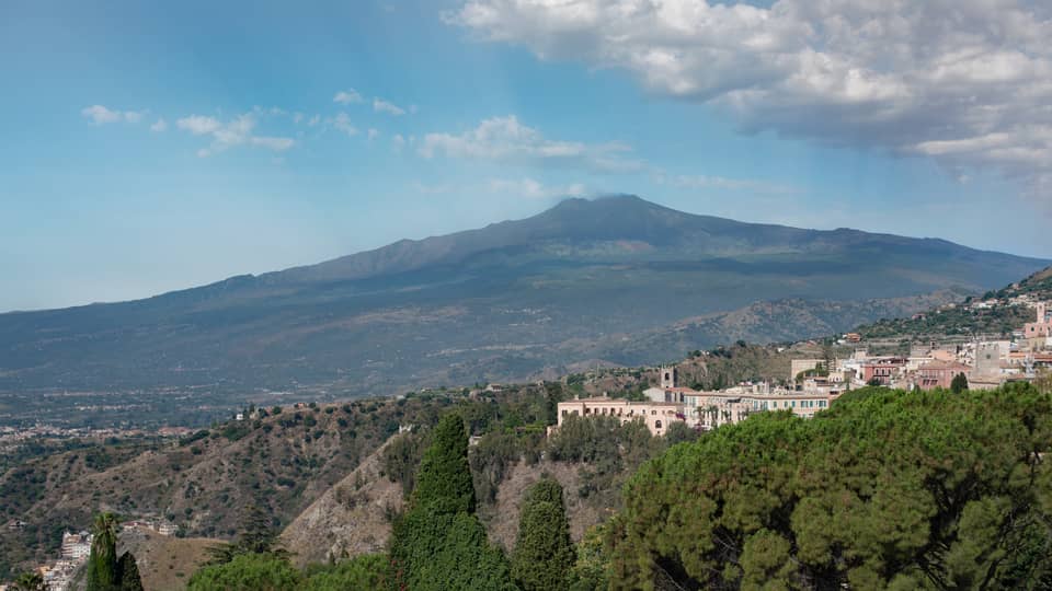 Hillside buildings surrounded by trees, Mount Etna and blue sky in backdrop
