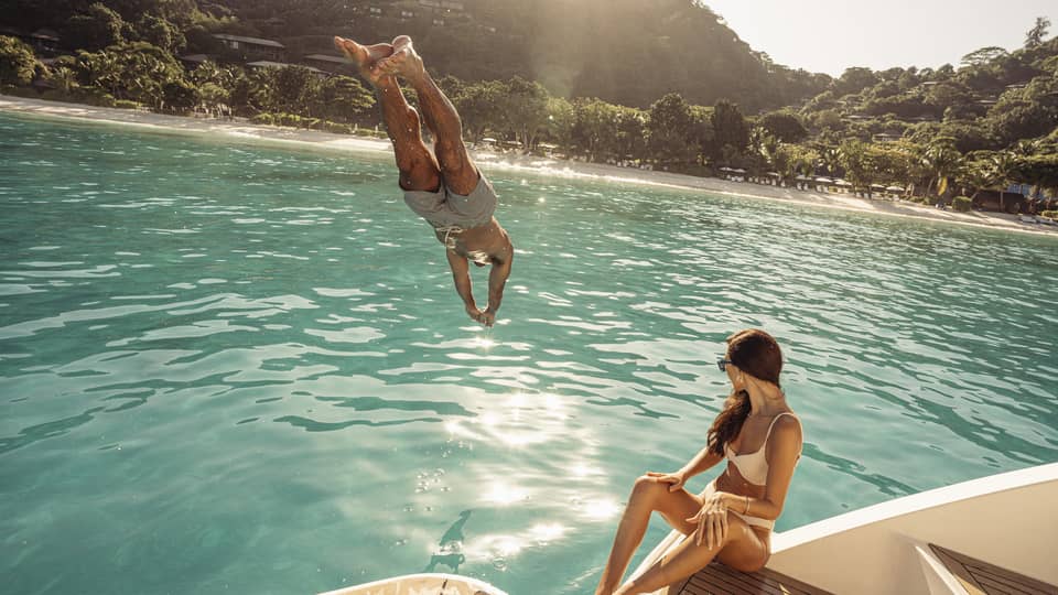 Person diving into blue-green water off a boat deck, where a person in swimsuit is sitting