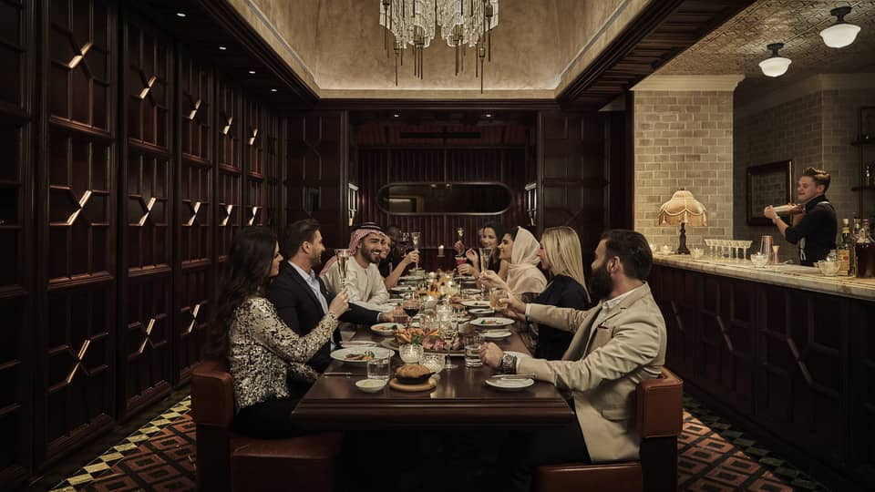 Ten people sit at a long, dark-wood rectangular table in a dark-wood panelled, speakeasy-style room with a crystal chandelier overhead and a marble bar counter and bartender to the side