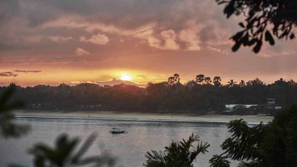 Sunrise over beach with trees and water, small boat 