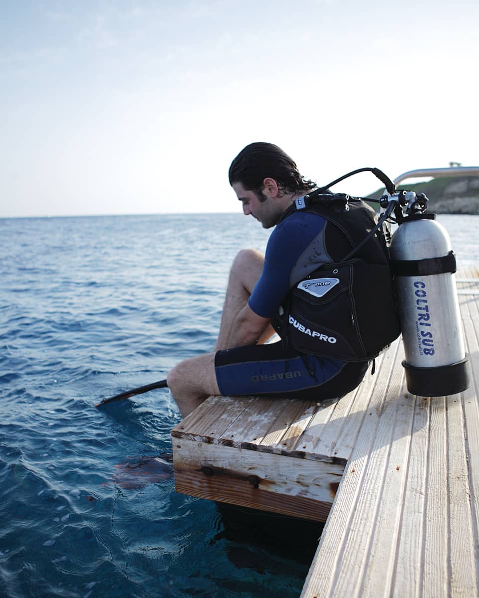 Man with wet hair wearing wetsuit, scuba diving tank sits on dock with feet in water 