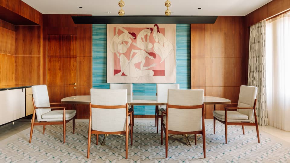 Dining room with a long table, six chairs, colorful abstract wall art and wood-paneled walls with natural light from large windows.