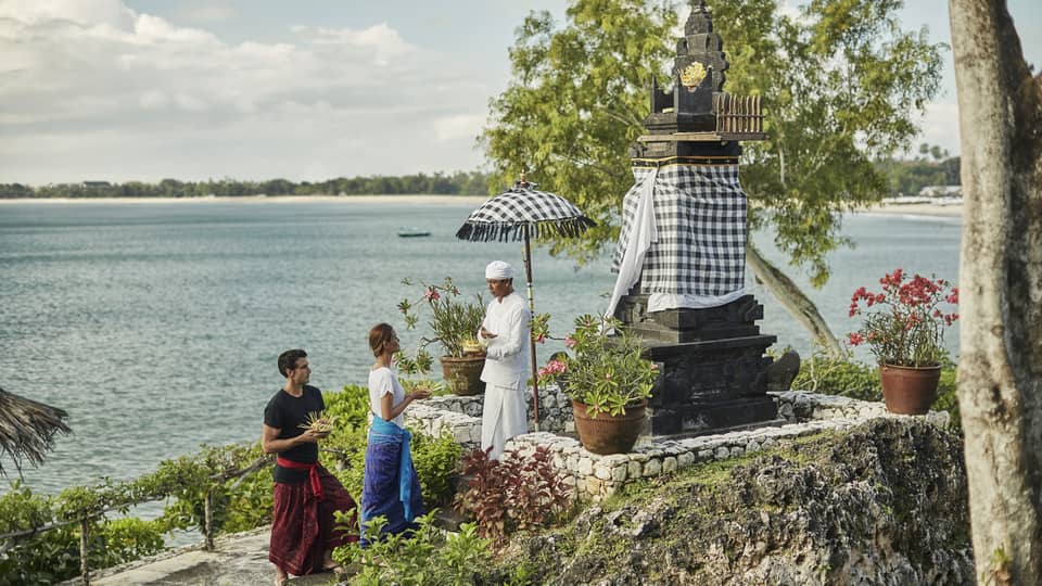 High Priest Aji Ngurah stands by shrine with black-and-white checkered umbrella, cloth, as man and woman make offering