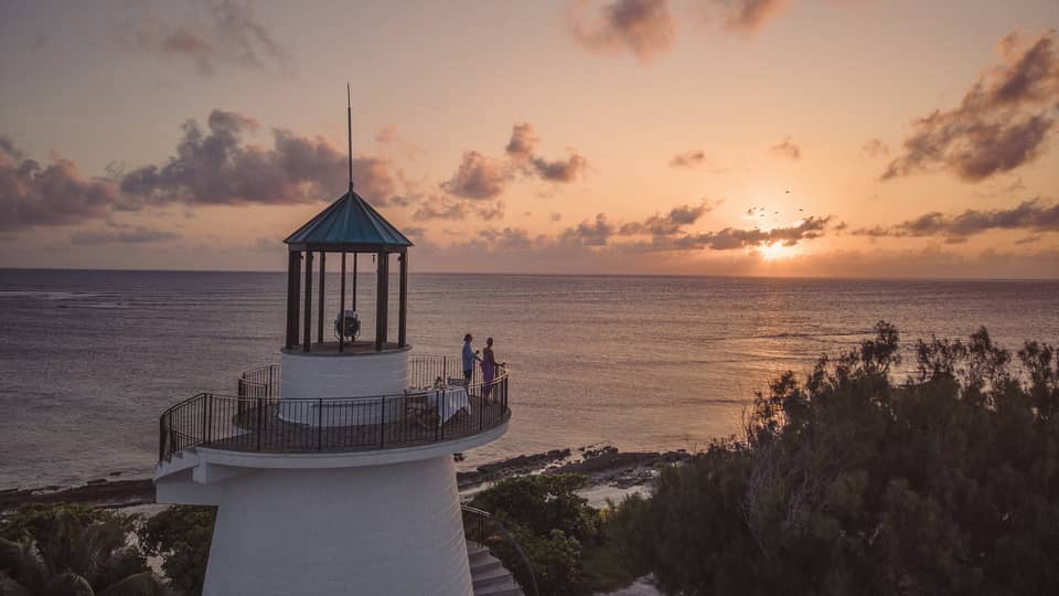 Long view of two people on a beach lighthouse balcony, enjoying the sunset over the ocean, distant silhouette of birds. 