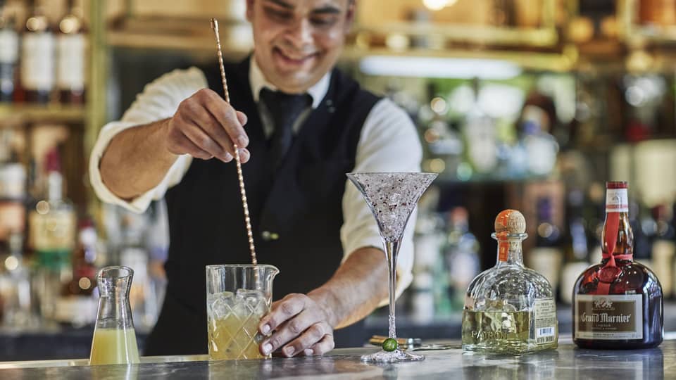 Bartender stirs cocktail with large swizzle stick on bar 