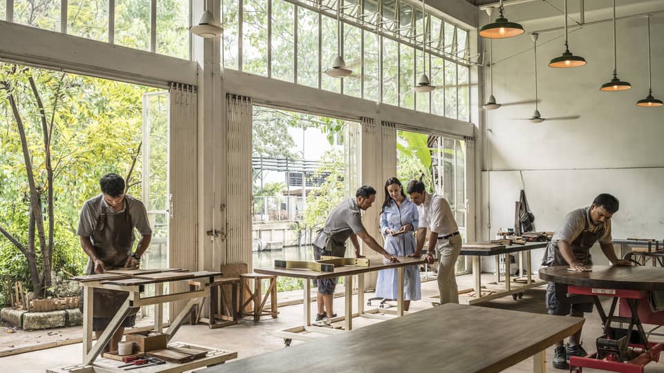 People working in a workshop with large windows, sanding and inspecting wooden furniture, while others collaborate at a table.