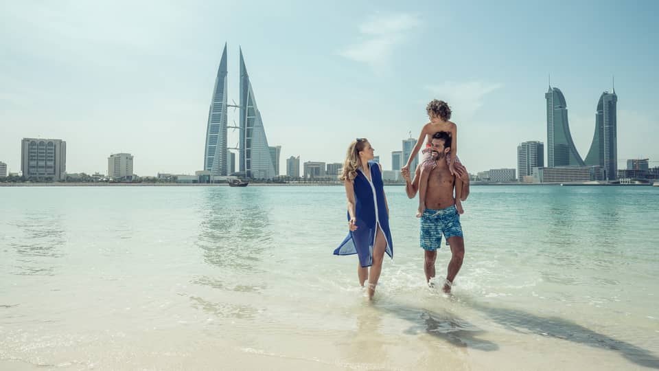 A man with a teenager on his shoulders walks with a woman through shallow ocean water with the city of Bahrain Bay in the background 