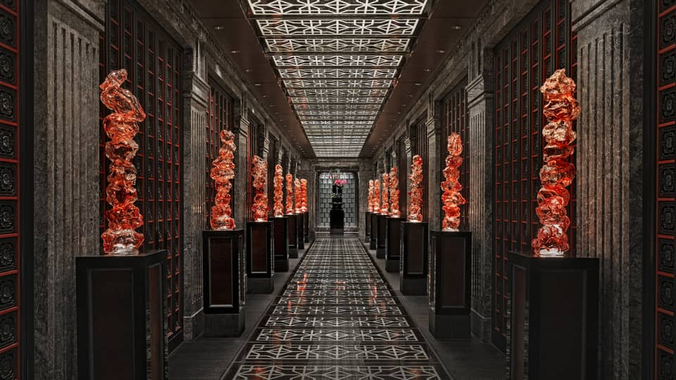 View down a long black-tiled hall lined with orange glass sculptures on pedestals, the geometric ceiling mirroring the floor.