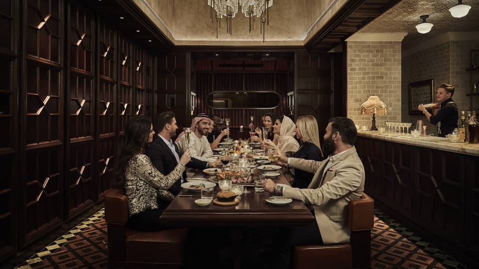 Ten people sit at a long, dark-wood rectangular table in a dark-wood panelled, speakeasy-style room with a crystal chandelier overhead and a marble bar counter and bartender to the side
