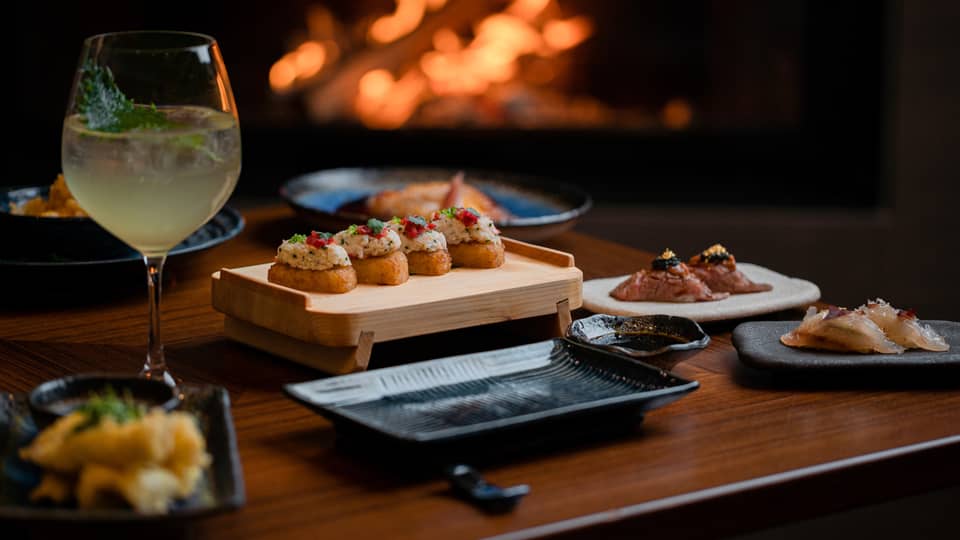 A cocktail and appetisers placed on a wooden table with a fireplace glowing in the background, creating a cosy dining atmosphere