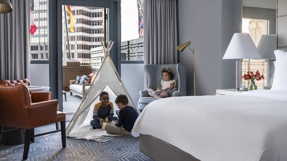 Children sitting in a tent and on a chair in a hotel room.