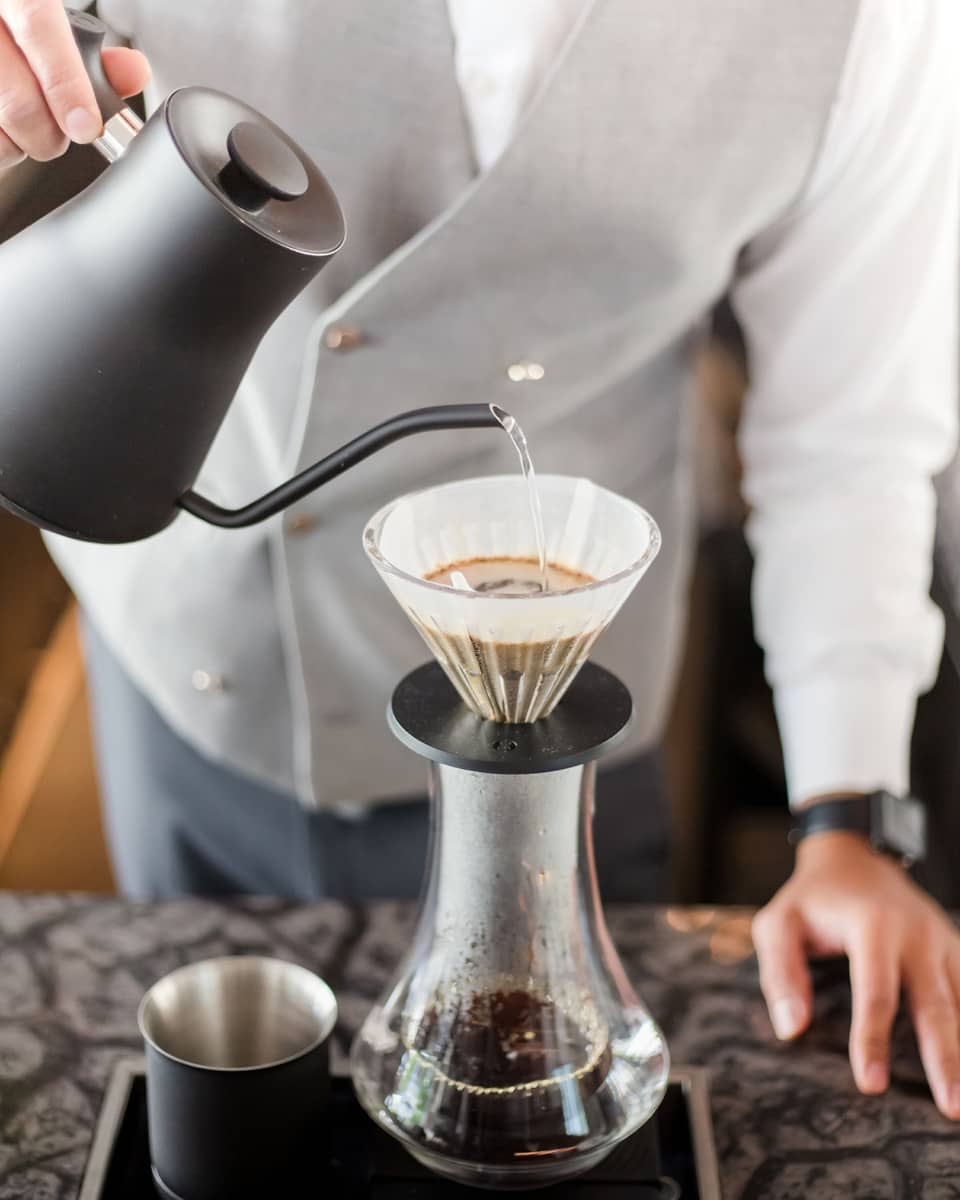 Barista holds black teapot, pours hot water over coffee in cone filter and glass jug on tray