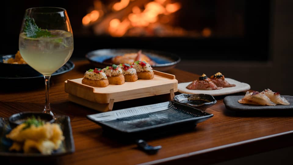 A cocktail and appetisers placed on a wooden table with a fireplace glowing in the background, creating a cosy dining atmosphere