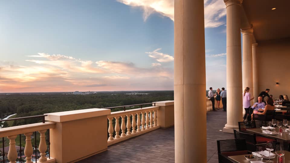 People dine at patio tables under tall pillars, balcony overlooking trees and sunset