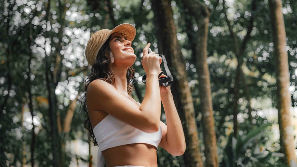 A woman stands in a tropical forest holding a camera, looking up