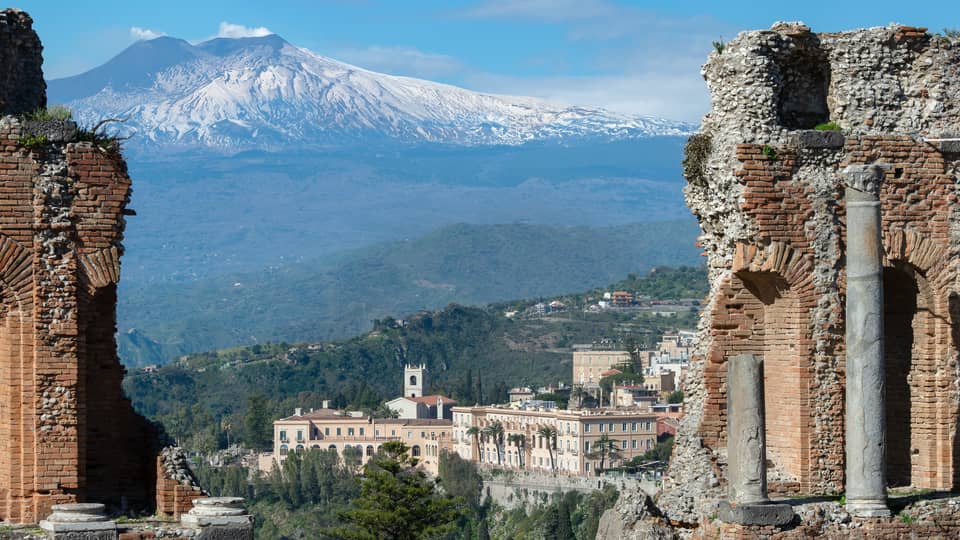 View of ancient stone ruins with a distant town and snow-capped mountains in the background.