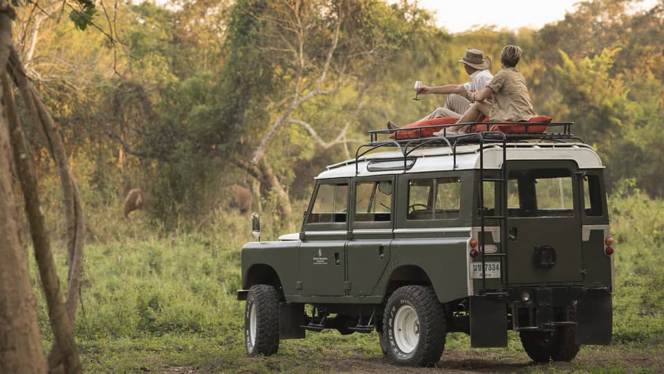 A couple sits on the roof of a Land Rover in the Bush, drinking a cocktail