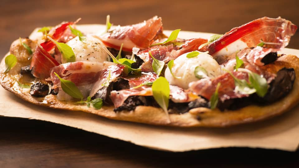 Close-up of thin crust Italian-style pizza topped with thinly-shaved cured meat, herbs, cheese
