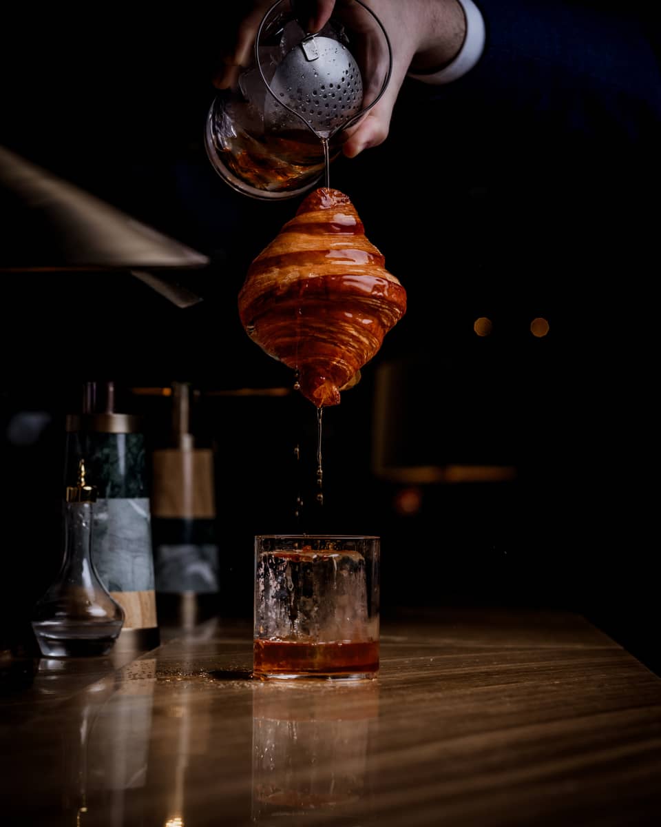 Bartender's hand pours a beverage out of a mixing glass onto a suspended croissant as it drips down into a rocks glass below