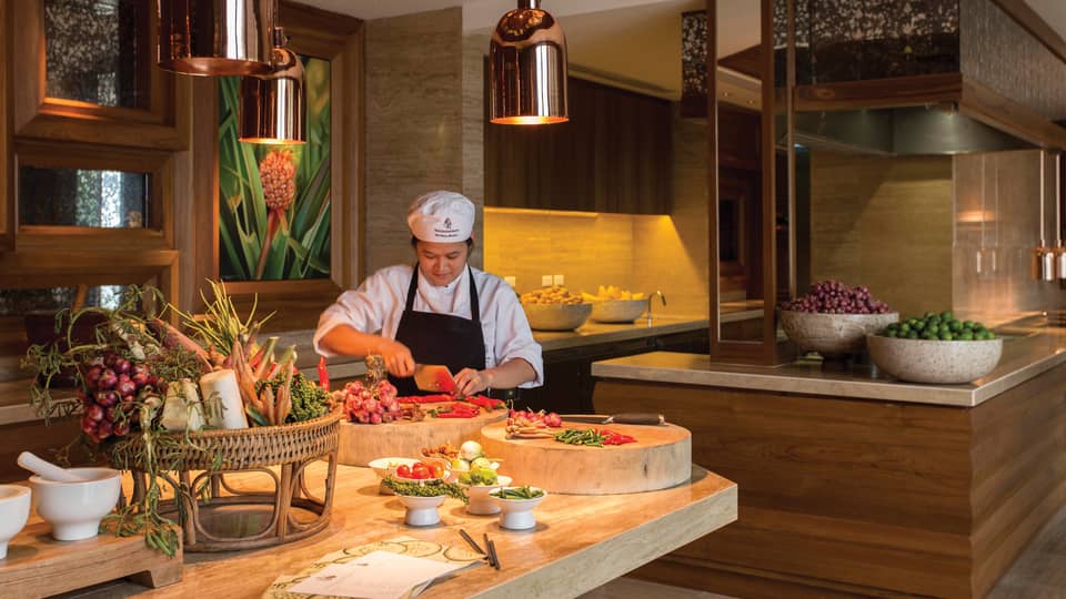 A chef chops vegetables on a large butcher block amid a vibrant array of fresh produce in a luxurious, warm-hued kitchen.