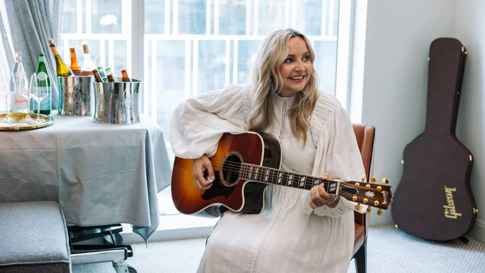 A woman with long blonde hair in a white dress plays an acoustic guitar while seated in a modern room with large windows, smiling and looking to the side.,,A woman with long blonde hair in a white dress plays an acoustic guitar while seated in a modern room with large windows, smiling and looking to the side.