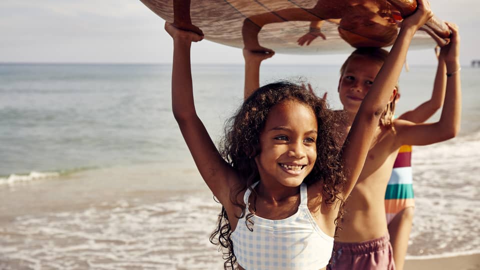 Three young children carry a surfboard on the beach, smiling and enjoying the sunny day. The ocean is in the background.