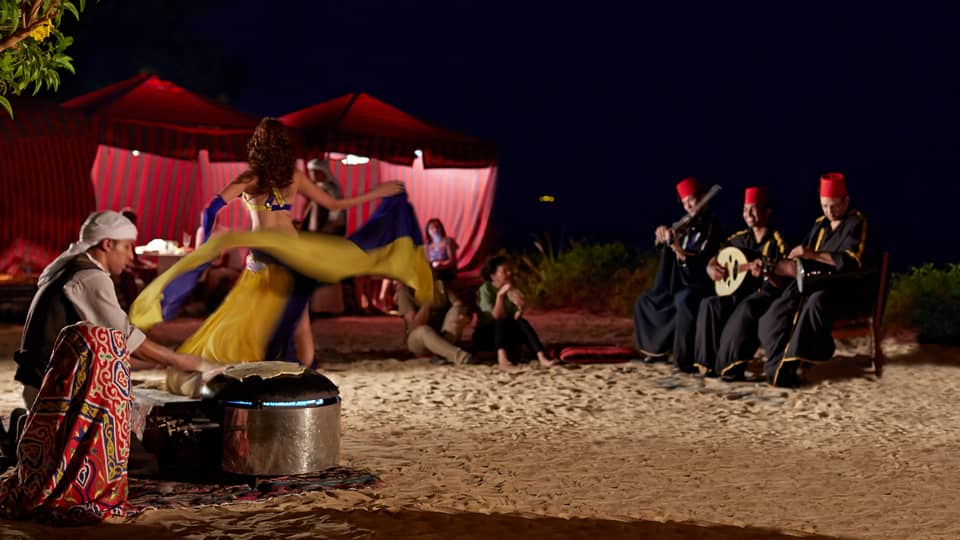At night, a dancer performs outdoors for guests, swirling their skirt to music played by three smiling, seated musicians.