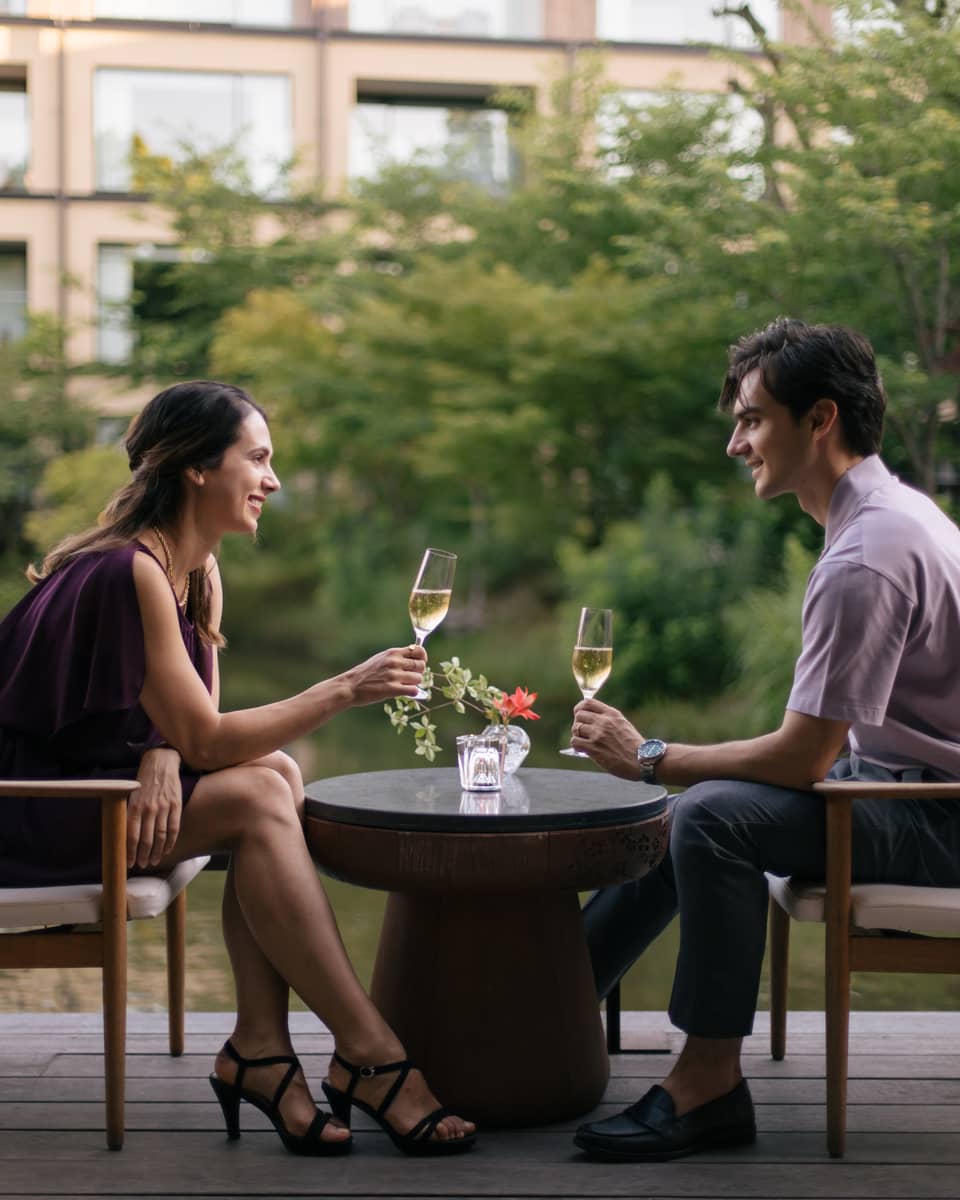 Two people sitting outdoors, toasting with champagne glasses at a small table with a flower vase