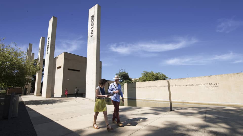 Man and woman walk on path past white pillars, wall with Nelson Mandela quote