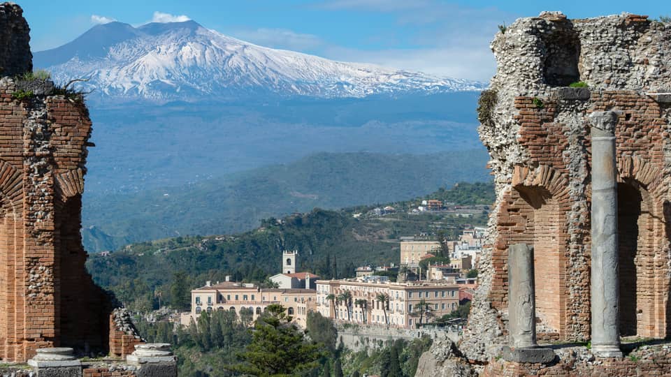 View of ancient stone ruins with a distant town and snow-capped mountains in the background.