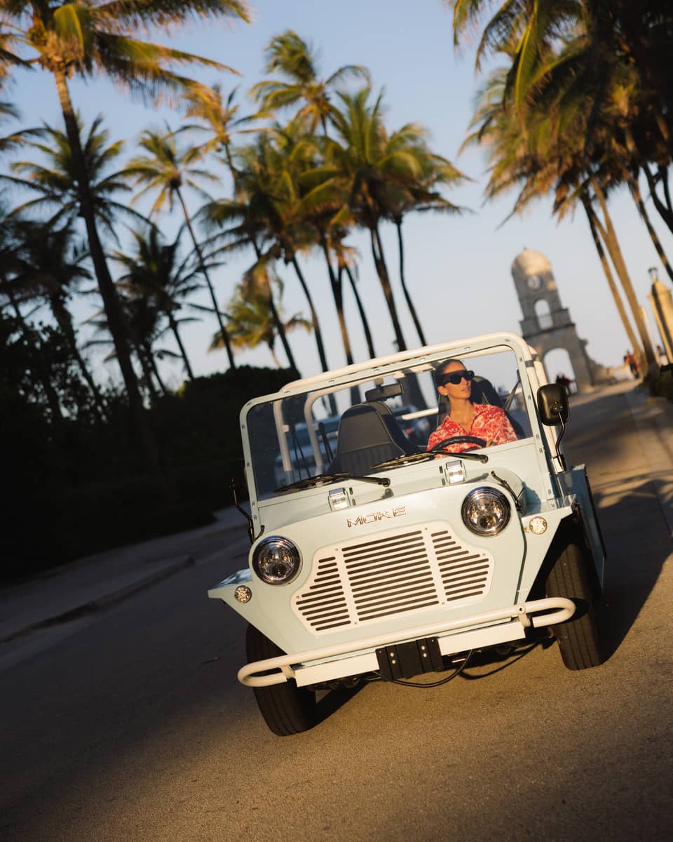 Guest wearing a red-and-white tropical shirt rides in a white MOKE along a palm tree?lined street at dusk
